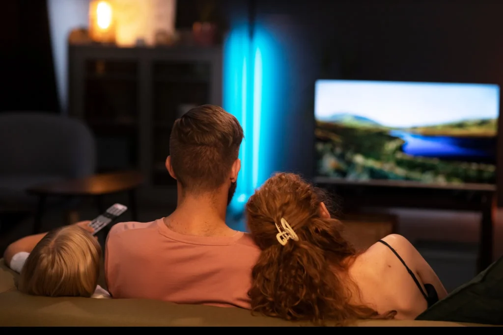 Family sits on a sofa watching a TV screen displaying a landscape, cozy living room scene at night.