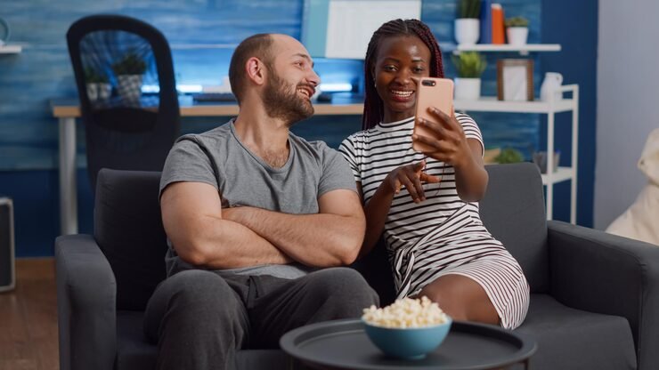 Man and woman sit on a dark sofa in a cozy living room, smiling at a phone the woman holds up for both to see, with a bowl of popcorn on a nearby table.